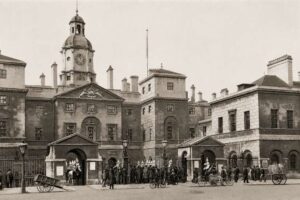 Household Cavalry Museum - London - Historische Aufnahme (1904): Eine alte Sepia-Fotografie des Horse Guards Gebäudes mit Passanten in historischer Kleidung, Pferdekutschen und Handkarren auf der Straße