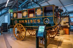 London Transport Museum - London - Pferdeomnibus: Der berühmte dunkelgrüne Shillibeer-Omnibus von 1829 mit großen gelben Holzrädern und bunten Plaketten mit Fahrtzielen