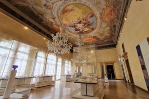 Museo del Vetro - Murano - Ausstellungsraum: Ein prunkvoller Saal mit historischer Deckenmalerei, Kronleuchtern und Glasvitrinen