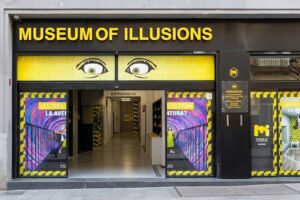 Museum of Illusions Madrid - Eingangsbereich: Frontale Aufnahme des Eingangs des Museums mit dem gelb-schwarzen Logo