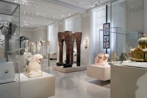 Victoria and Albert Museum (V&A) - South Kensington - London - Ausstellung Faiths and Empires: Ein moderner Raum mit antiken Säulenkapitellen, Steinskulpturen und religiöser Kunst in beleuchteten Glasvitrinen